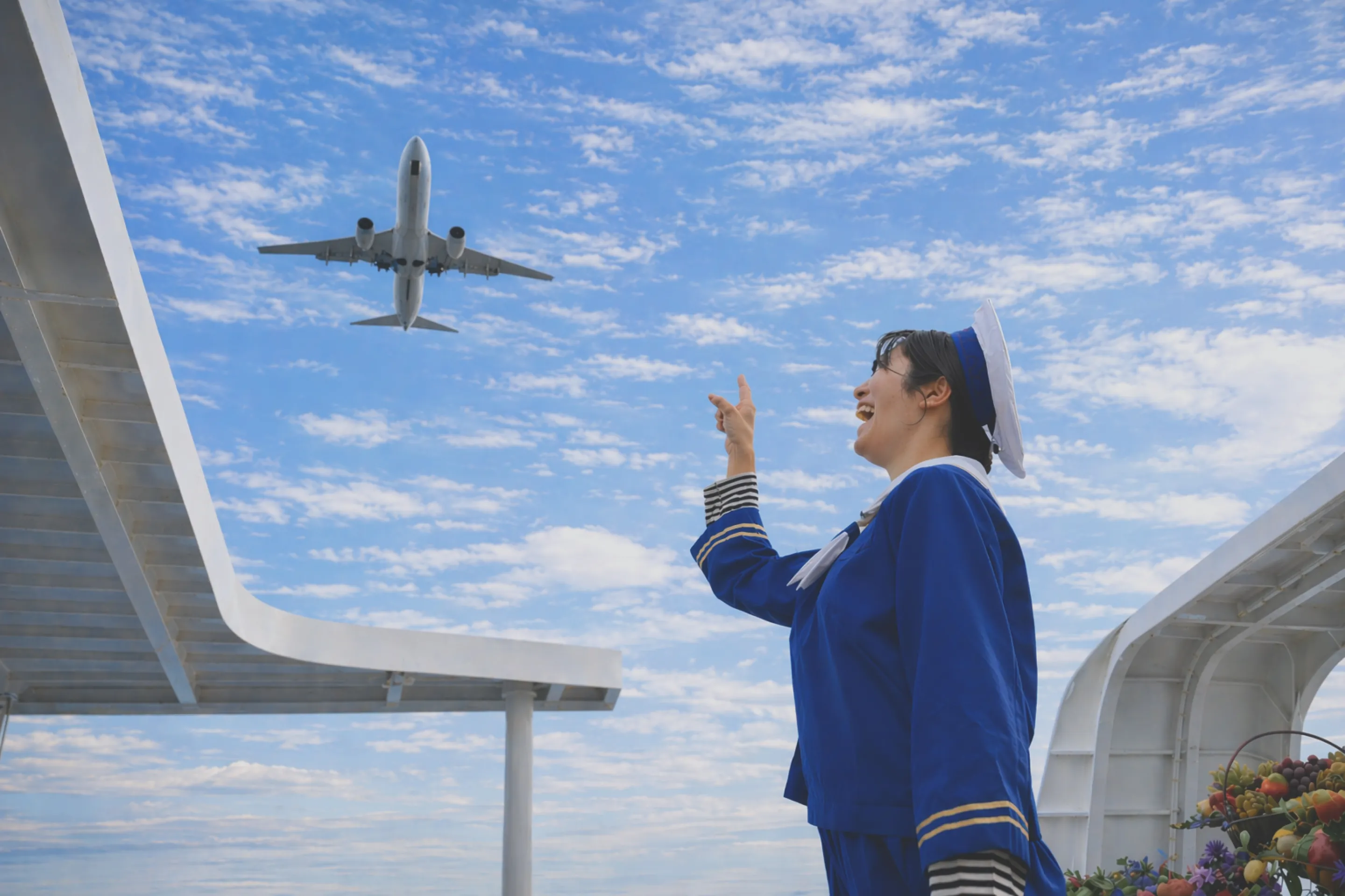 空飛ぶ飛行機を指差す、セーラ服とセーラ帽を身につけたモデルの写真